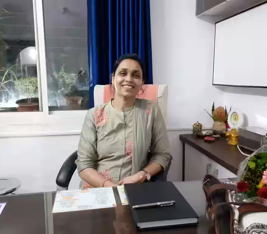 Dr. Shraddha Bihani smiling at her desk in a well-lit office with blue curtains and floral decor.